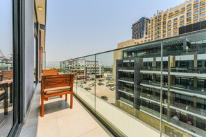 A balcony with a wooden bench, a glass railing, and a view of a parking lot and buildings. - HAVN - Modern 2 Bed in Oxford 212, JVC (Dubai)