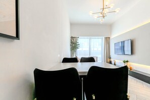 A modern dining area with a long table, black chairs, a mounted TV, and a large window with curtains. - HAVN - Modern 2 Bed in Oxford 212, JVC (Dubai)