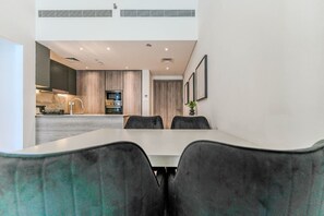 A modern dining area with a long table and chairs, a kitchen in the background, and a ceiling with recessed lighting. - HAVN - Modern 2 Bed in Oxford 212, JVC (Dubai)