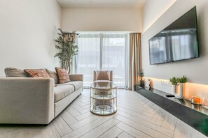 Uma sala de estar moderna com sofá, mesa de centro de vidro, televisão na parede e uma grande janela com cortinas.
