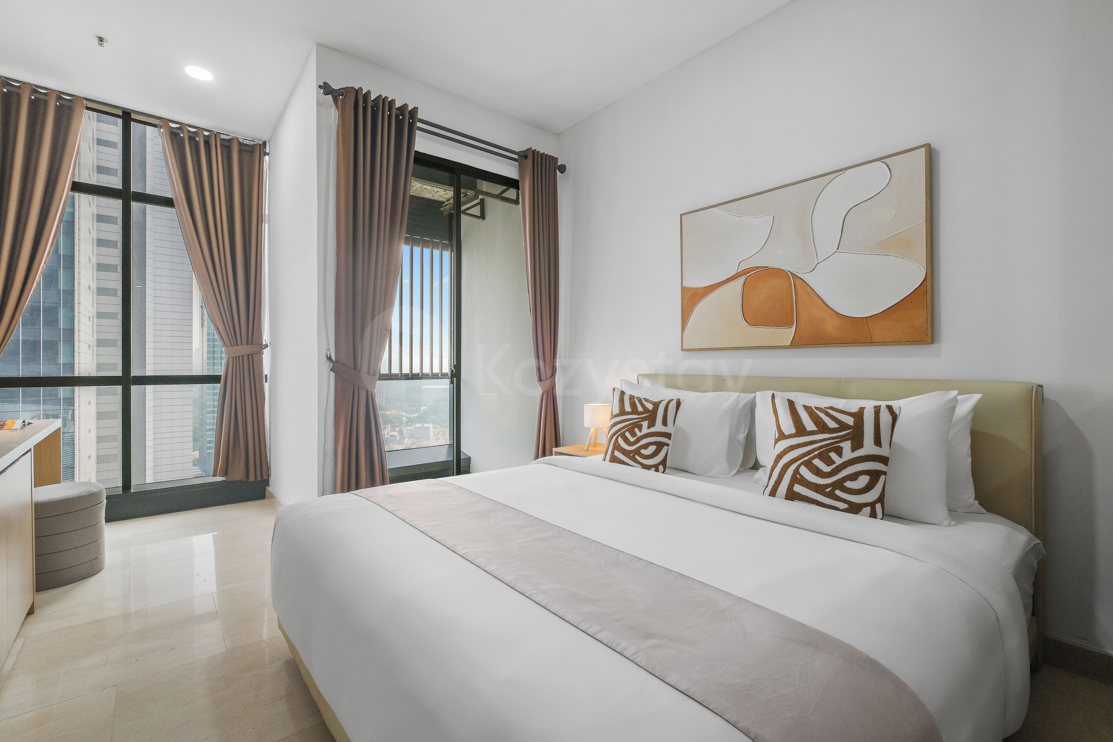 A modern hotel room with a large bed, a desk, and a view of the city through floor-to-ceiling windows.