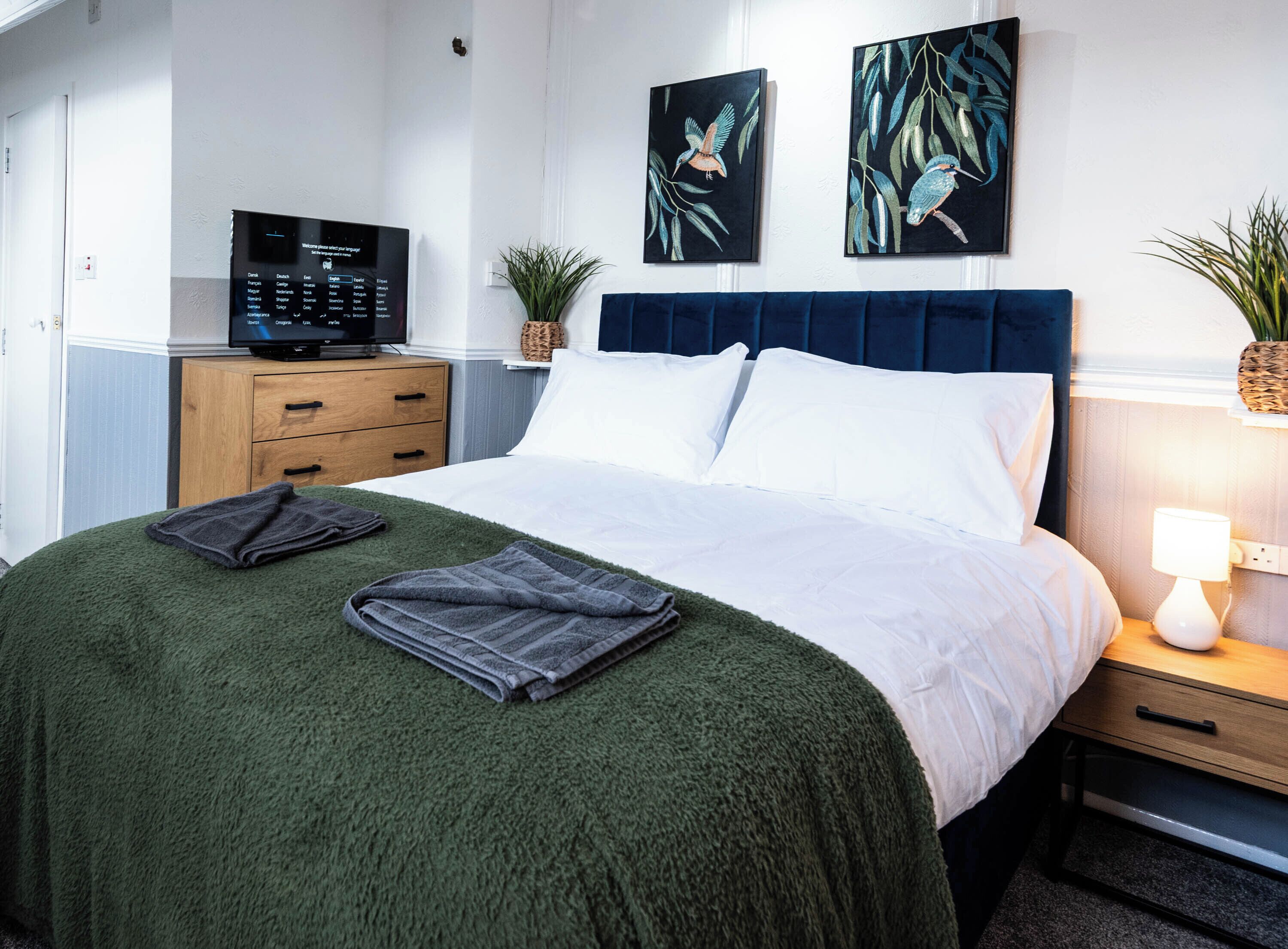 A neatly made bed with a green blanket, a bedside lamp, and two paintings on the wall.