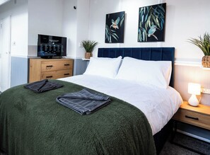 Ein ordentlich bezogenes Bett mit grüner Decke, einer Nachttischlampe und zwei Bildern an der Wand.