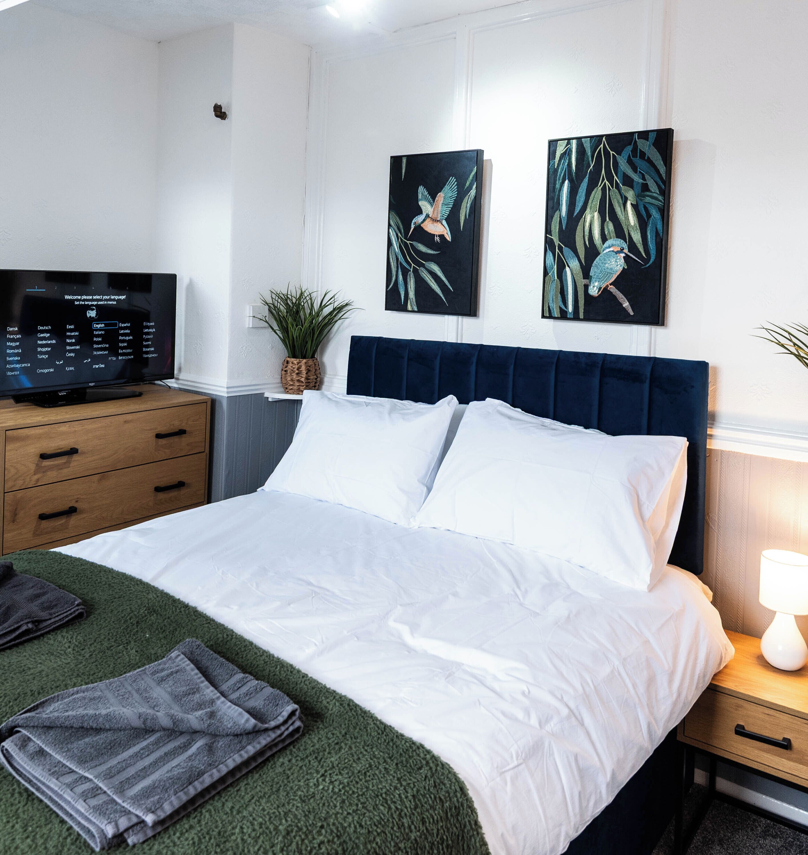A neatly made bed with white linens, a dark blue headboard, and a television on a wooden dresser to the left.