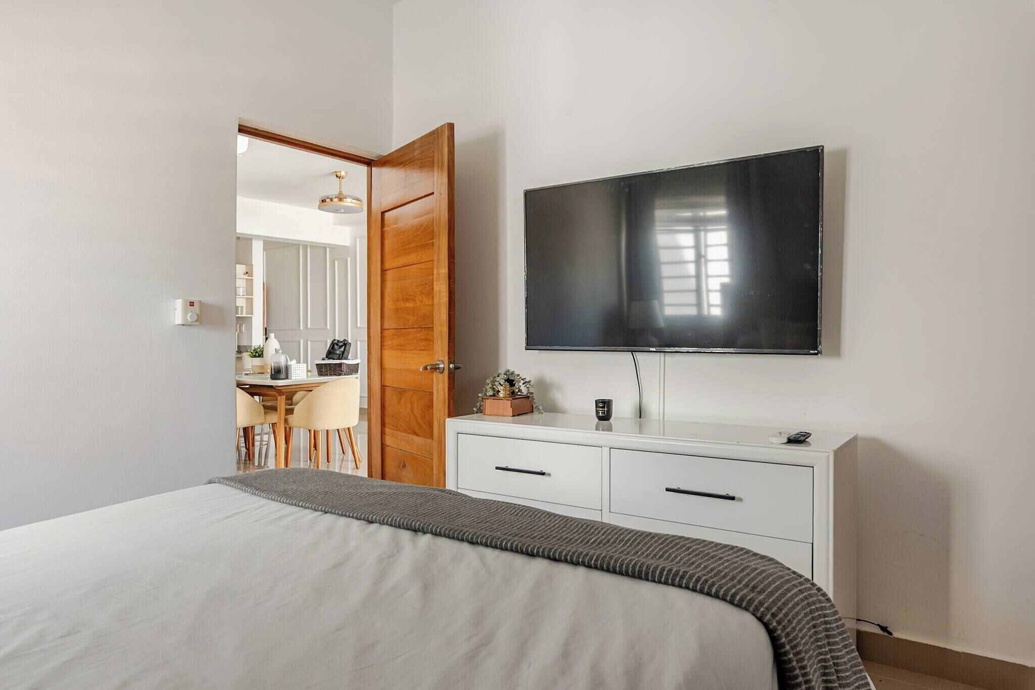Een slaapkamer met een bed, een tv die aan de muur is bevestigd, een houten deur en een bureau met stoelen op de achtergrond.