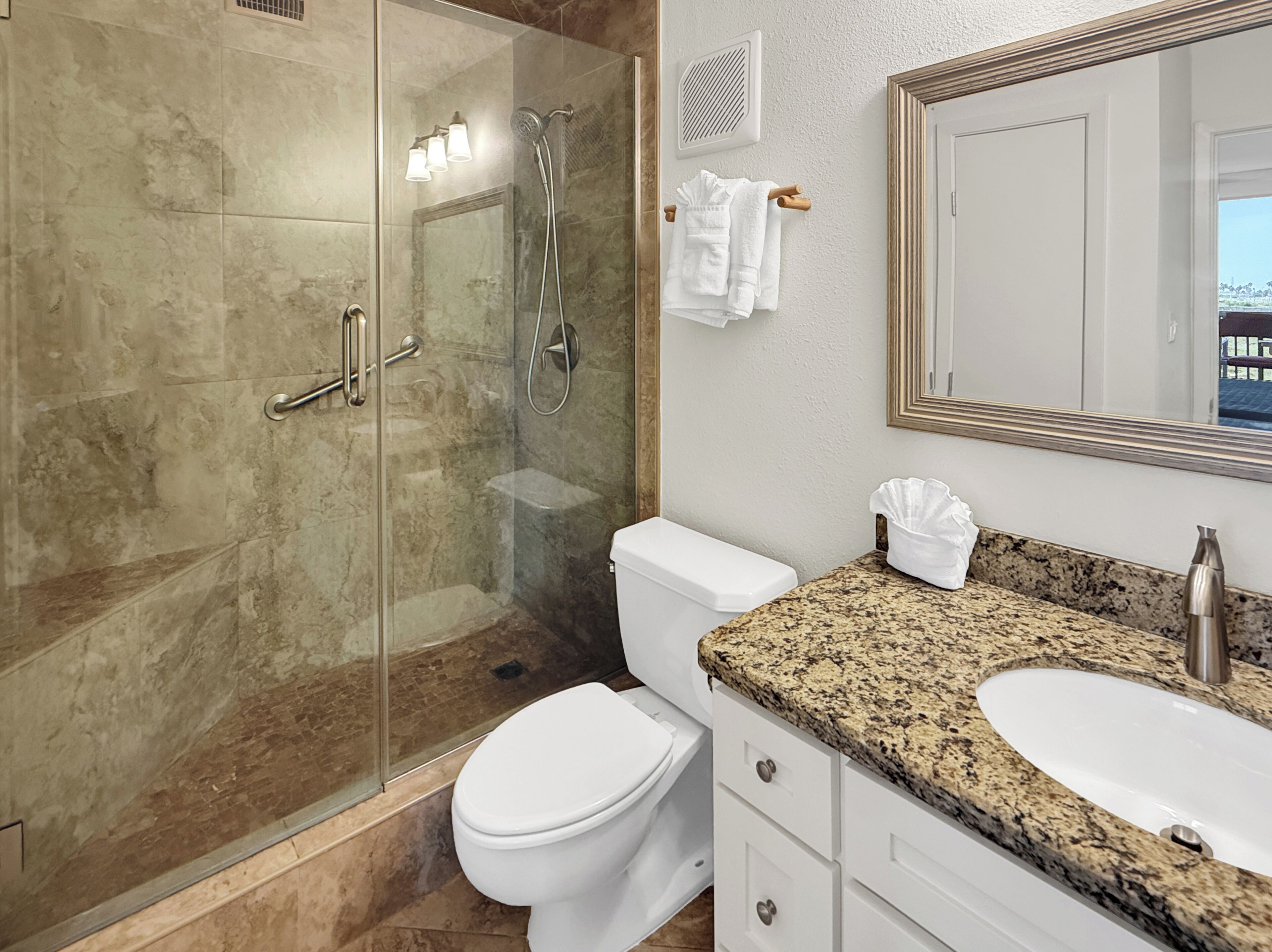 A modern bathroom with a glass-enclosed shower, a white toilet, a granite countertop, and a large mirror.