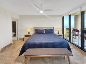 Quarto com cama, mesas de cabeceira, um banquinho, ventilador de teto e vista para o exterior.