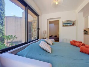 A bedroom with a large window, a bed with blue bedding, a nightstand, a lamp, and a painting on the wall. - Les Sources by Interhome (Draguignan)