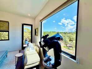 Ein Zimmer mit einem Teleskop, einer Couch, einem kleinen Tisch und einem großen Fenster mit Blick auf Bäume.
