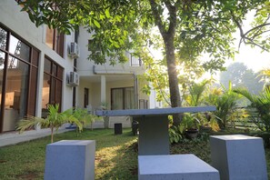 A courtyard with a concrete bench, a small palm tree, and a building with large windows and air conditioning units. - Superb 4-bedroom house with AC in charming Kandy (Kegalle)