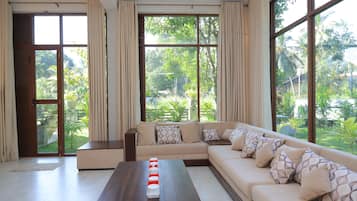 Un salon spacieux avec un grand canapé d’angle, une table basse en bois et une vue sur un jardin luxuriant grâce à de grandes fenêtres.