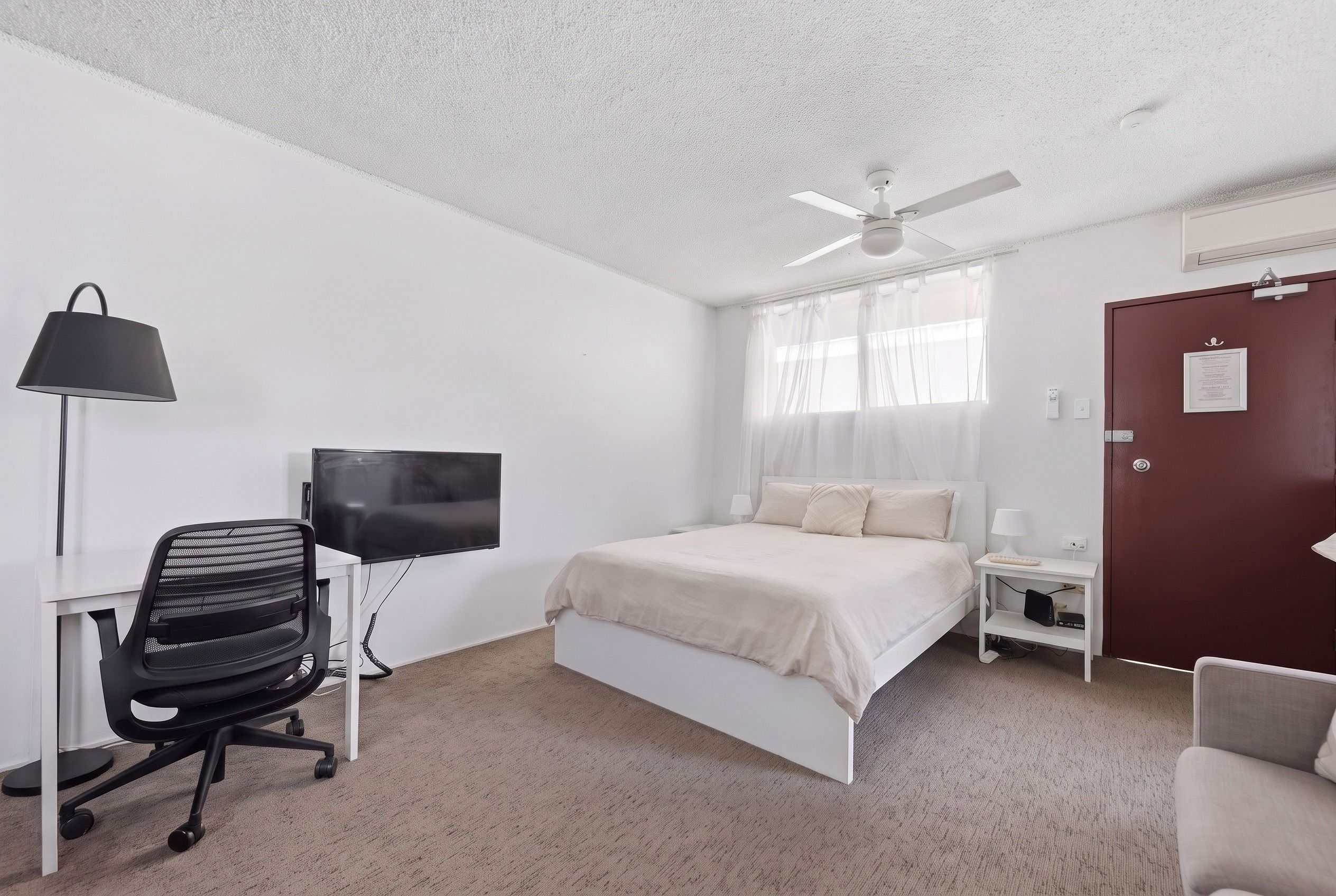 Une chambre d’hôtel comprenant un lit, un bureau avec une chaise, une télévision, un ventilateur de plafond et une porte avec une pancarte.