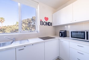A modern kitchen with white cabinets, a black coffee maker, a microwave, and a window with a view of trees. - Bondi Beach Studio | 2 Min Walk to Beach, Sleeps 4, Work-From-Beach Desk (Bondi Beach)
