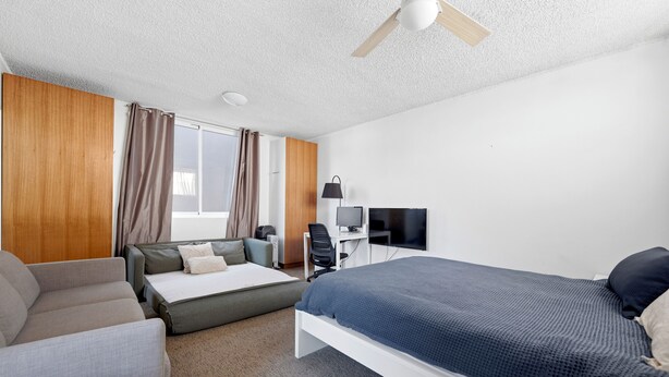 Ett hotellrum med en säng, en soffa, ett skrivbord med en dator, en TV och en takfläkt.