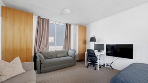 Ett hotellrum med en soffa, ett skrivbord med en dator och en datorskärm, en tv och ett fönster med gardiner.