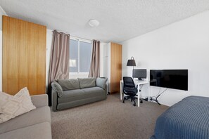 Ein Hotelzimmer mit einer Couch, einem Schreibtisch mit Computer und Monitor, einem Fernseher und einem Fenster mit Vorhängen.