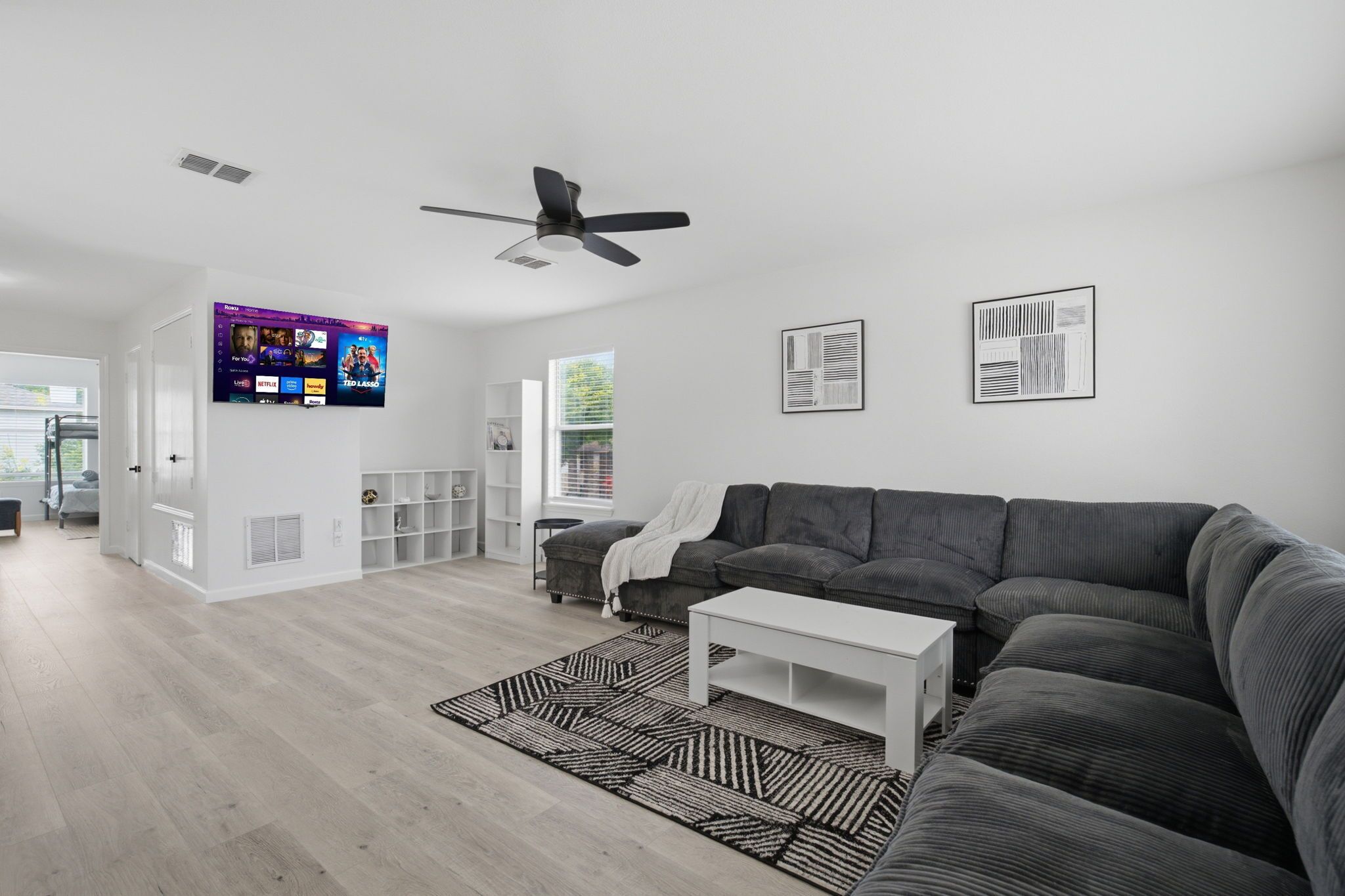 A modern living room with a grey sectional sofa, a white coffee table, a black and white rug, a ceiling fan, and a mounted TV displaying streaming services.
