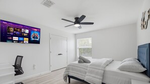 Una camera da letto con un letto, una scrivania, una sedia, una ventola a soffitto, una TV montata a parete e una finestra con tende.