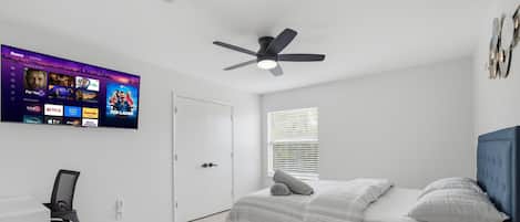 Quarto com cama, escrivaninha, cadeira, ventoinha no teto, TV fixada na parede e janela com persianas.