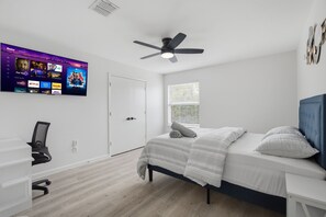 Ein Schlafzimmer mit einem Bett, einem Schreibtisch, einem Stuhl, einem Deckenventilator, einem an der Wand montierten Fernseher und einem Fenster mit Jalousien.