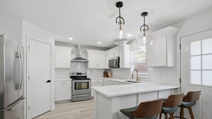 Una cucina moderna con mobili bianchi, un'isola centrale con sgabelli da bar, elettrodomestici in acciaio inossidabile e illuminazione a sospensione.