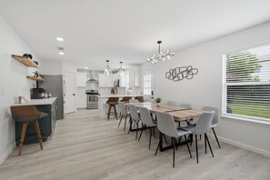 A modern kitchen with white cabinets, a dining area with a wooden table and chairs, and a large window with blinds. - New Luxury & Spacious 5BR/3BA Retreat–Sleeps 16 – Prime location & Fully Stocked (McKinney)