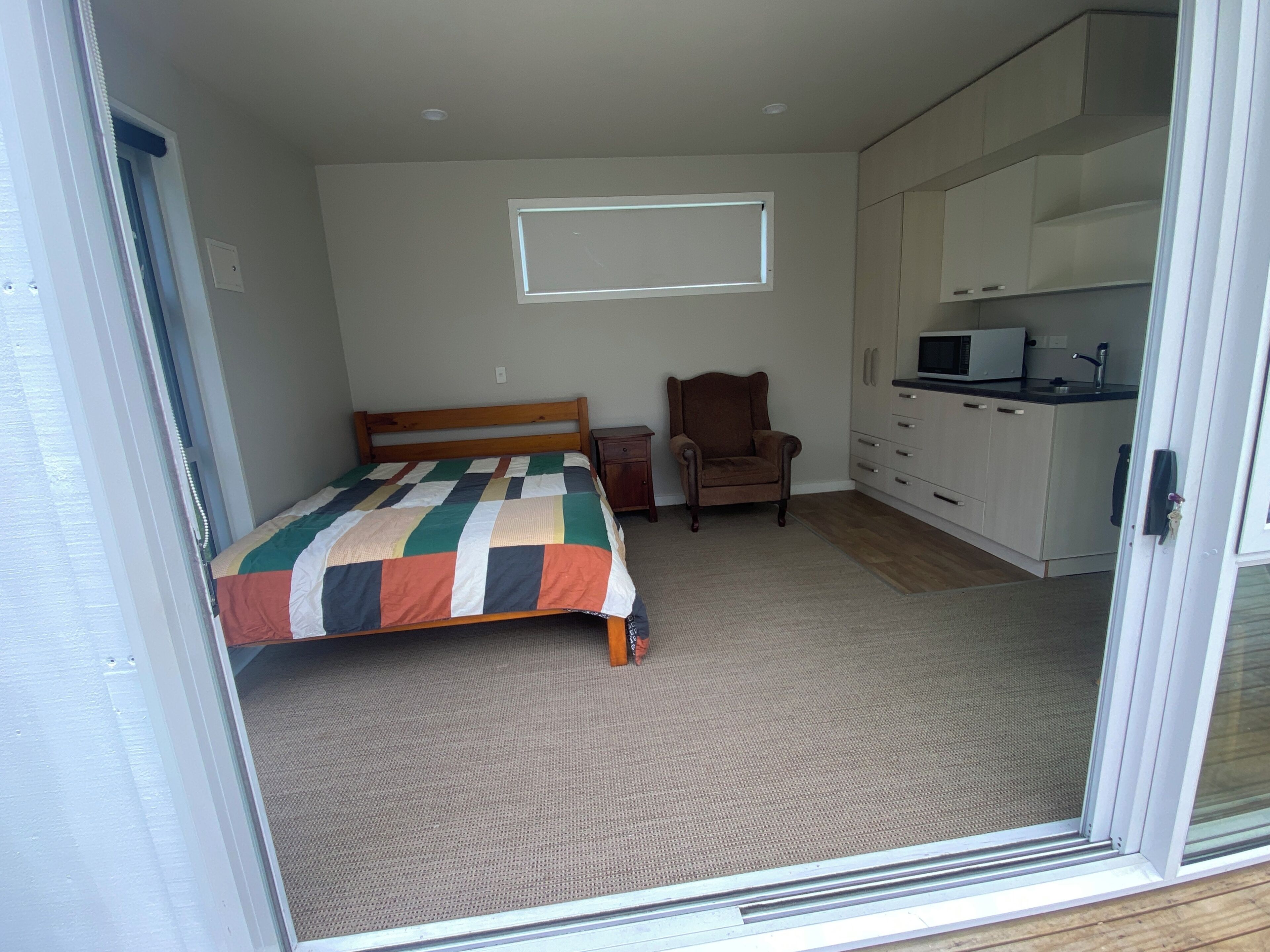 Een slaapkamer met een bed, een stoel, een tafeltje, een raam en een kitchenette.