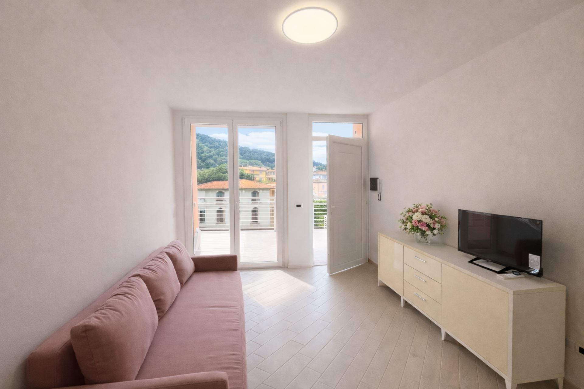 Una habitación moderna con sofá, televisión sobre un mueble y una puerta que da a un balcón con vistas a edificios.