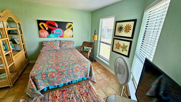 A bedroom with a bed, a rattan chair, a fan, and a window with blinds.
