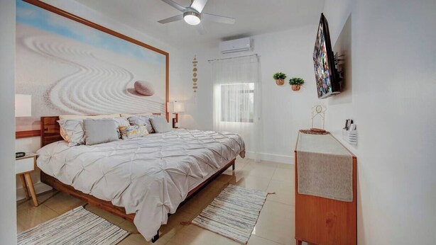 A bedroom with a bed, bedside tables, a ceiling fan, a wall-mounted TV, a small plant, and a window with curtains. - Glamorous 2Bed/2Bath/2K- 5★ DT Location-♛Hot Tub (Fort Lauderdale)