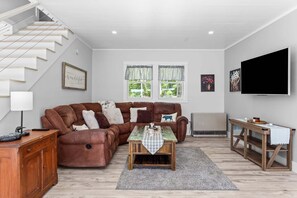 A living room with a brown leather sofa, a wooden coffee table, a TV on the wall, and a staircase. - Toddy View - NEW! Waterfront Home on Toddy Pond (Blue Hill)