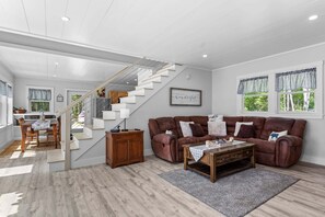 A modern living room with a brown leather sofa, a wooden coffee table, and a staircase leading to an upper level. - Toddy View - NEW! Waterfront Home on Toddy Pond (Blue Hill)