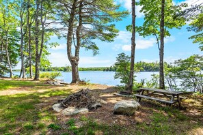 A lakeside campsite with a picnic table and a fire pit surrounded by trees. - Toddy View - NEW! Waterfront Home on Toddy Pond (Blue Hill)