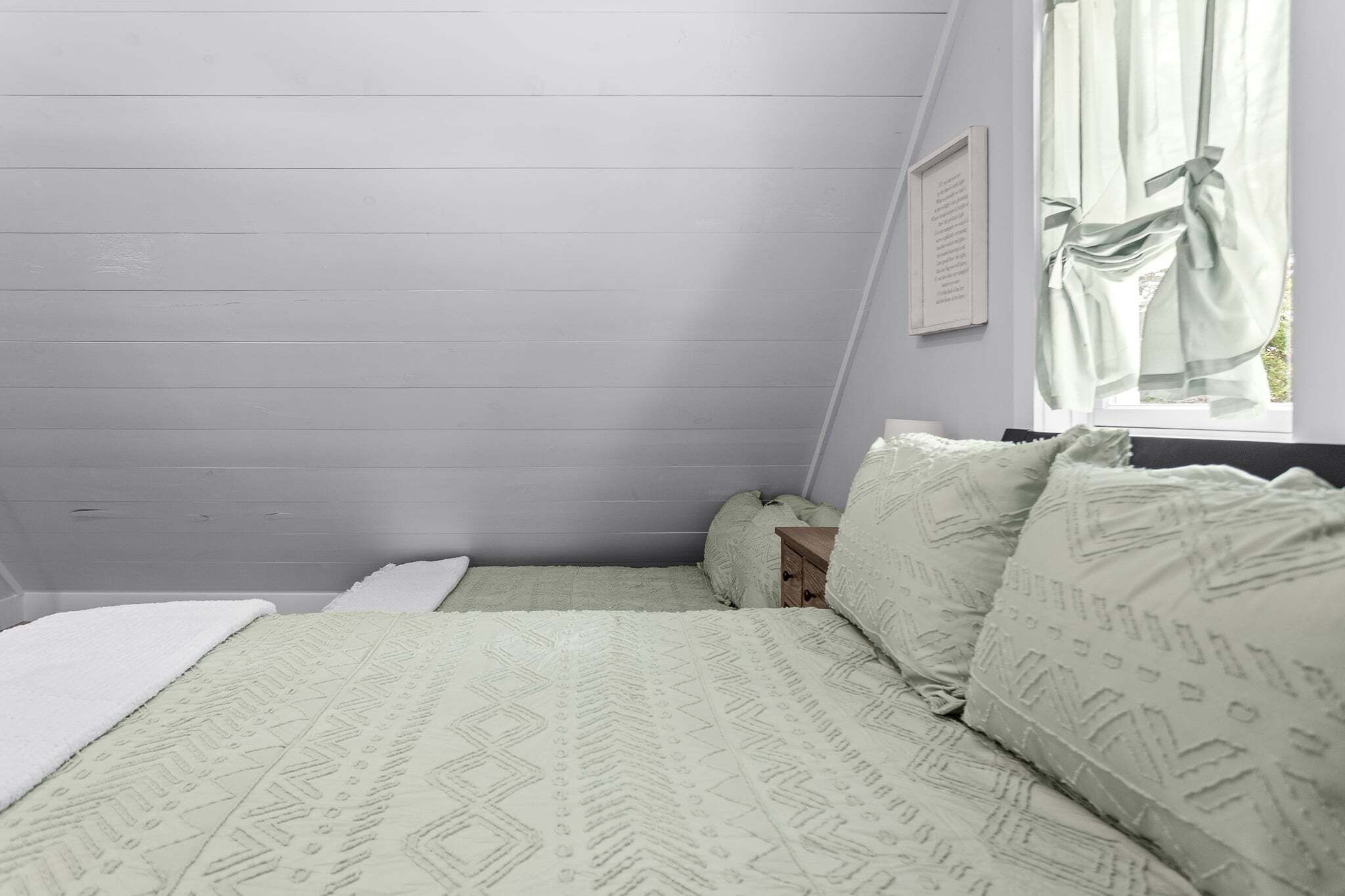 A bedroom with a sloped ceiling, a bed with patterned bedding, a bedside table, and a window with curtains.