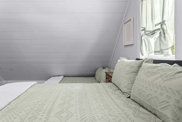 A bedroom with a sloped ceiling, a bed with patterned bedding, a bedside table, and a window with curtains. - Toddy View - NEW! Waterfront Home on Toddy Pond (Blue Hill)