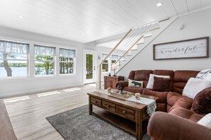 A spacious living room with a brown leather sofa, a wooden coffee table, and a staircase leading to another level. - Toddy View - NEW! Waterfront Home on Toddy Pond (Blue Hill)
