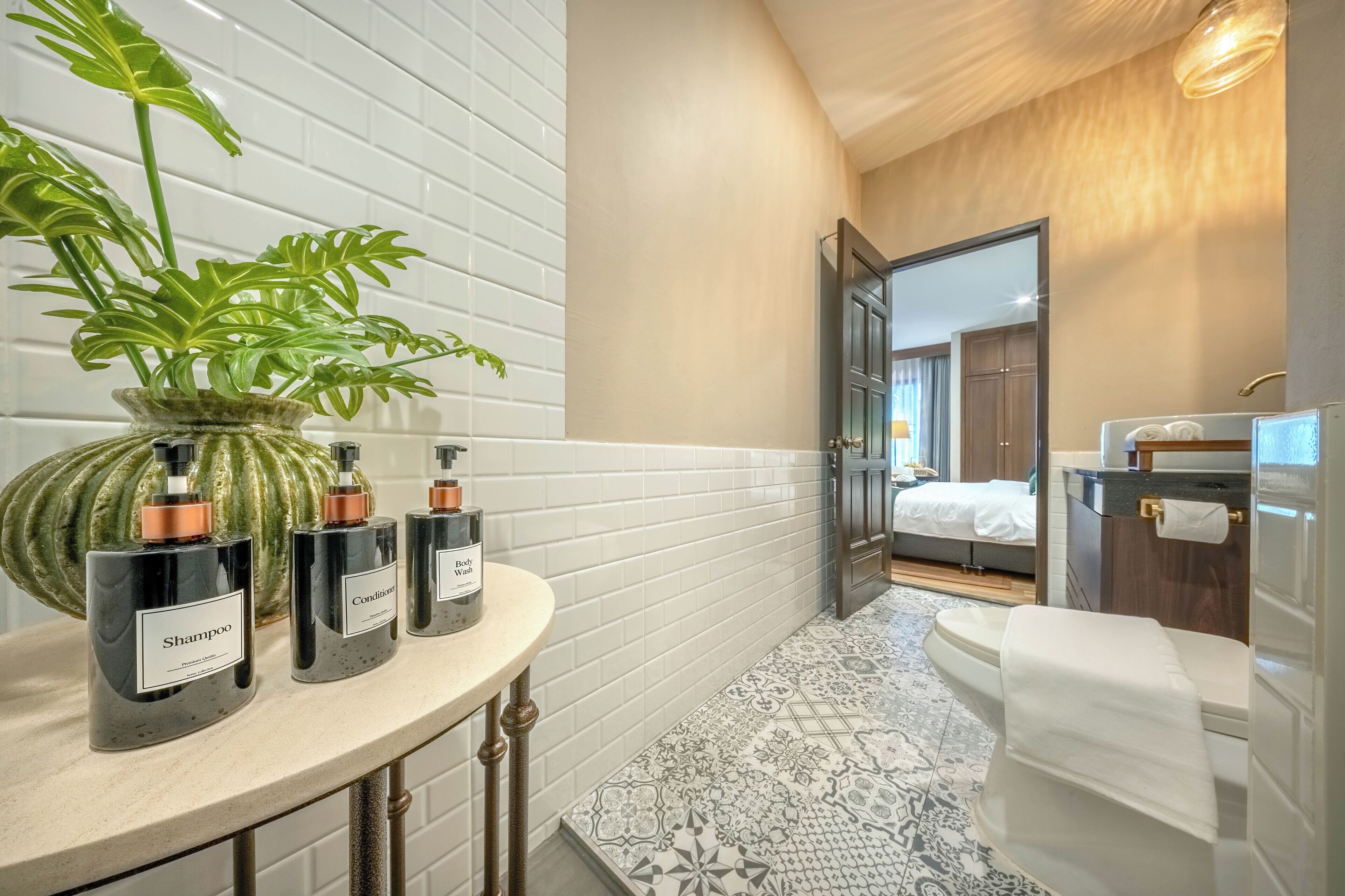 Ein Badezimmer mit weiß gefliester Wand, einer großen Topfpflanze, drei Flaschen Shampoo und Conditioner, einer Toilette und einem Spiegel.