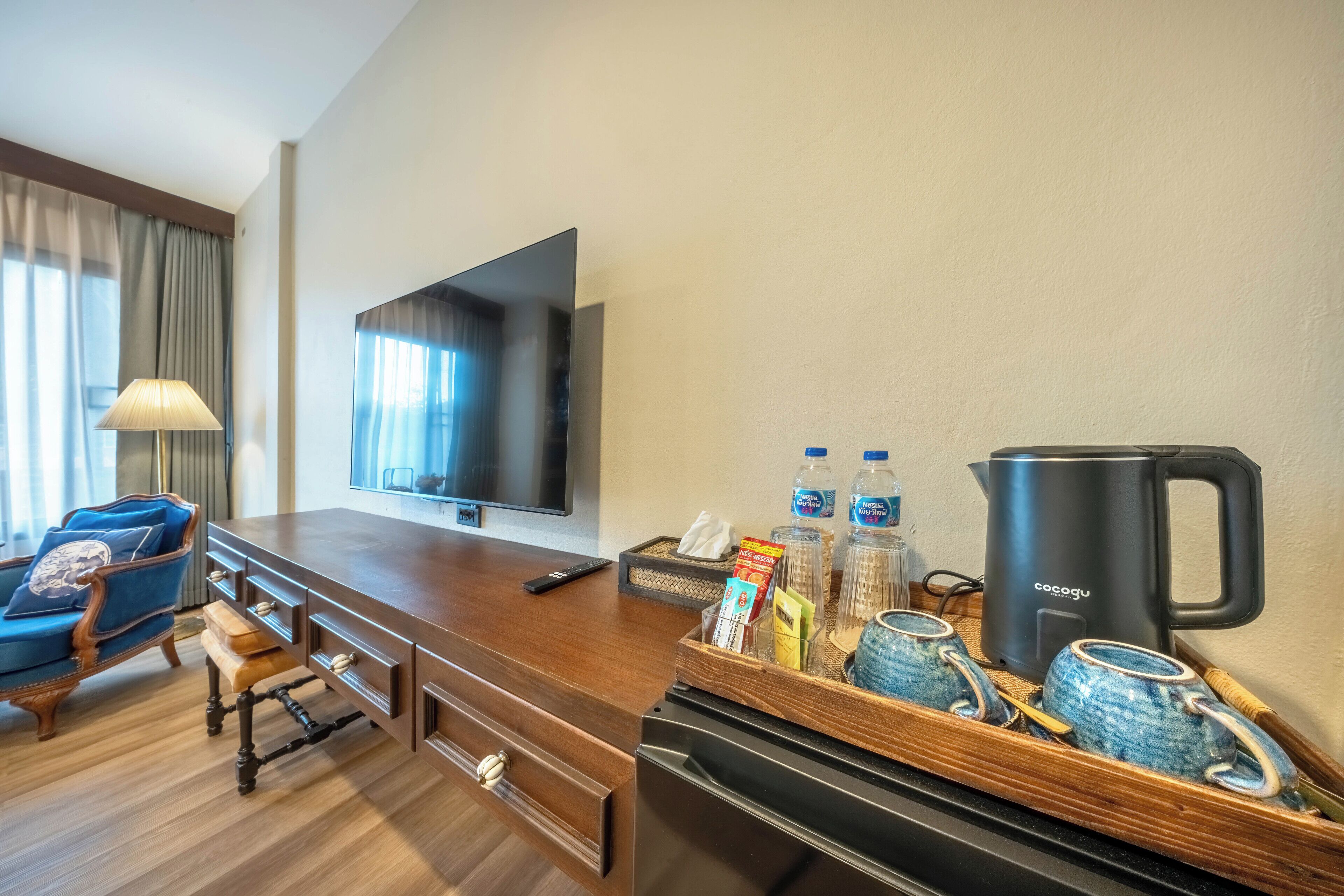 Ein Hotelzimmer mit einem Holztisch, einem Fernseher, einer Minibar, einer Kaffeemaschine und einem blauen Sessel.