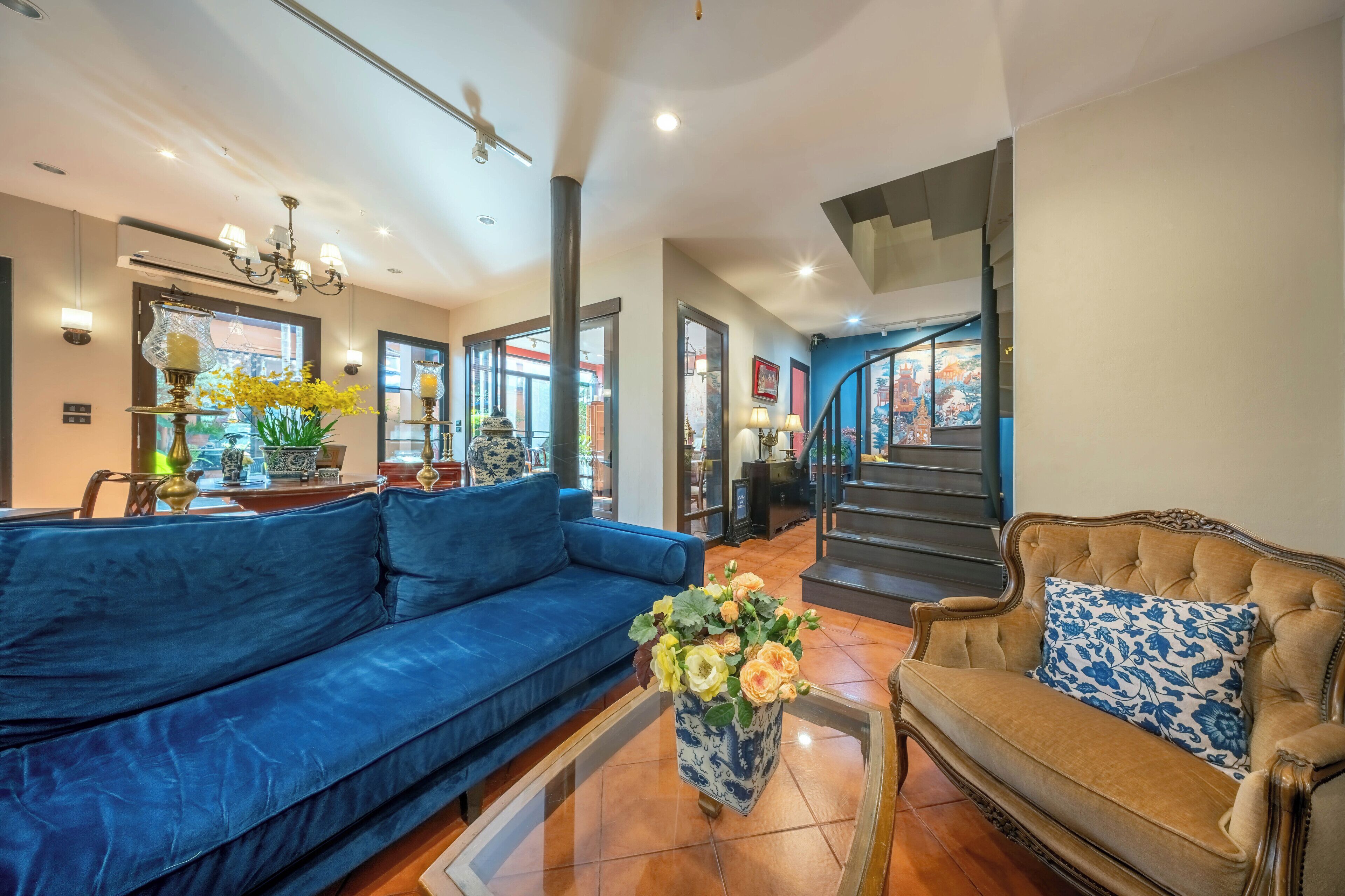 A spacious living area with a blue sofa, a brown sofa with a blue pillow, and a coffee table with flowers. There is a staircase leading to another level, and a large window with a view of a building.