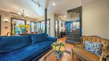 A spacious living area with a blue sofa, a brown sofa with a blue pillow, and a coffee table with flowers. There is a staircase leading to another level, and a large window with a view of a building.