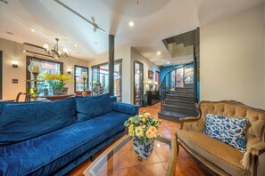 A spacious living area with a blue sofa, a brown sofa with a blue pillow, and a coffee table with flowers. There is a staircase leading to another level, and a large window with a view of a building. - Aphirom Hometel (Bangkok)