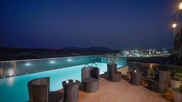 A rooftop pool area with wicker furniture and a city view at night.