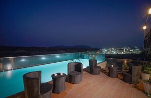 A rooftop pool area with wicker furniture and a city view at night.