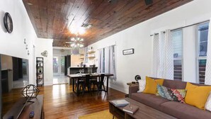 Un soggiorno moderno con soffitto in legno, un divano con cuscini gialli, un'area pranzo con tavolo e sedie, e una cucina sullo sfondo.