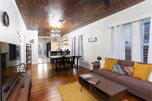 Ein modernes Wohnzimmer mit Holzboden, einer Couch mit gelben Kissen, einem Essbereich mit Tisch und Stühlen sowie einer Küche im Hintergrund.
