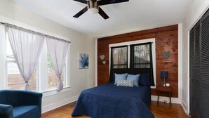 Una camera da letto con un letto, una sedia, una ventola a soffitto, una finestra con tende e un muro in legno.