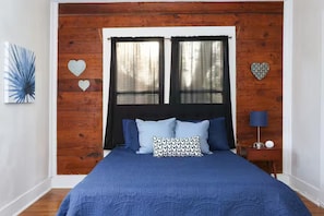 A bedroom with a wooden headboard, a bed with blue bedding, two windows with sheer curtains, a bedside table with a lamp, and decorative heart-shaped wall art. - Cozy Home 10 minutes to Riverwalk and Downtown (San Antonio)