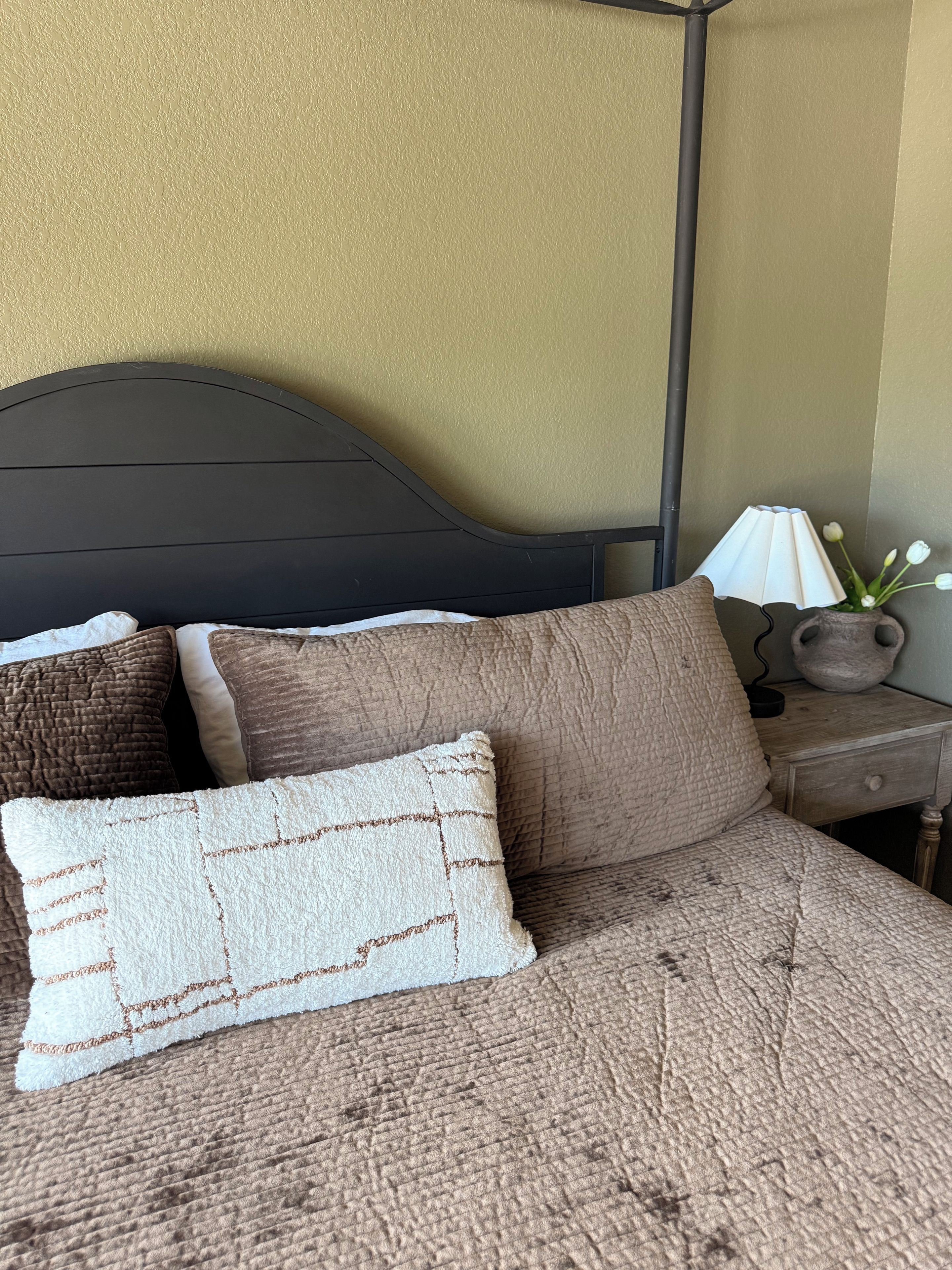 Una cama con cubrecama marrón texturizado, dos almohadas, cabecera oscura, lámpara de noche y un florero con flores sobre una mesita de noche de madera.