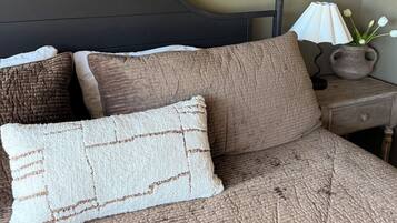 A bed with a textured brown cover, two pillows, a dark headboard, a bedside lamp, and a vase with flowers on a wooden nightstand.