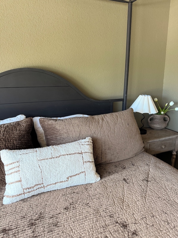 A bed with a textured brown cover, two pillows, a dark headboard, a bedside lamp, and a vase with flowers on a wooden nightstand.